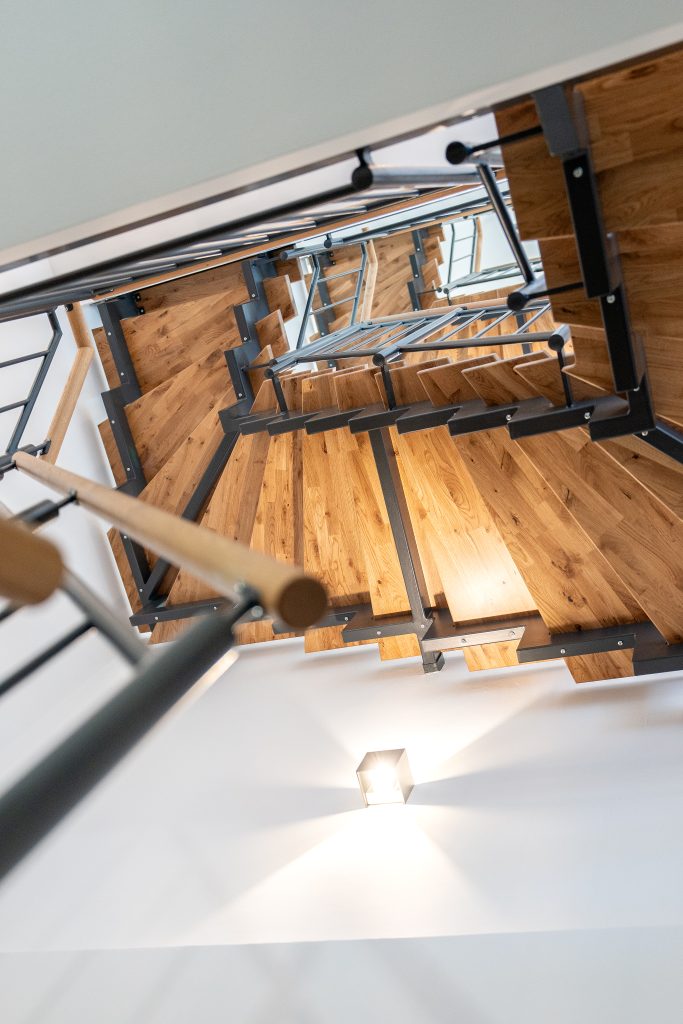 Blick vom Keller in das zweite Stockwerk durch die Holztreppe mit schwarzen Akzenten und hölzernem Handgriff.