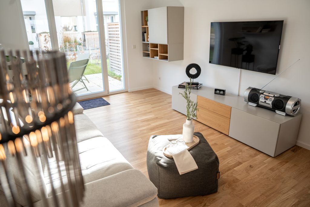 Das Wohnzimmer mit bodentiefen Fenstern und einer Glastür in den Garten. Im Raum stehen ein Sofa mit Beistelltisch. Der Boden ist aus Holz.